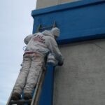 Man on a ladder painting blue trim using a brush on a commercial storage building