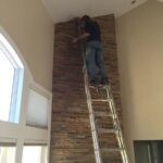 Man on a ladder painting a ceiling above a tall fireplace in a residential house