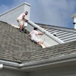 Two men working on a roof painting brick and trim using a sprayer and roller