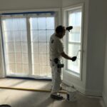 Man rolling white paint onto wood trim around interior window of a home