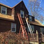 Painter on the ladder applying semi transparent stain dark brown to cedar siding on the back of a house