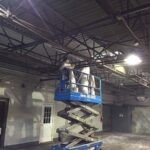 Metal steel deck ceiling in a factory being spray painted white by two painters in a scissor lift