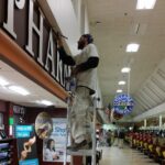 Painter standing on a ladder using a brush to apply maroon paint around large letters in a retail grocery store