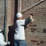 Tuckpointing being applied to a partially painted brick wall on an old commercial factory building by a painter on a step ladder