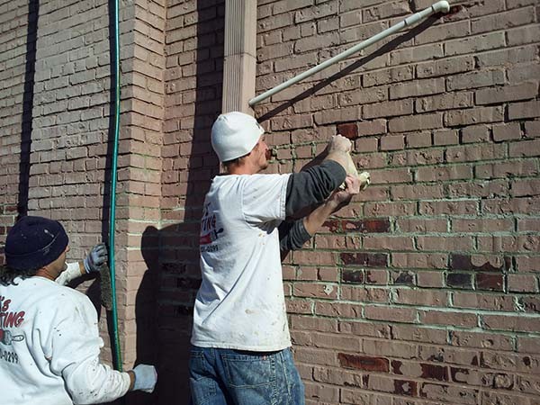 Tuckpointing being applied to a partially painted brick wall on an old commercial factory building by a painter on a step ladder