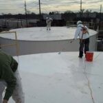 Three men painting the tops of two metal tanks white using rollers