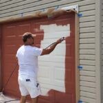 Man spraying red paint onto steel overhead garage door on a residential home