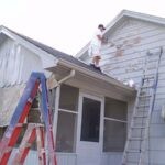 One painter on a roof painting wood siding with white paint and a roller, second man on a ladder painting trim and siding