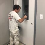 Men wearing DJ's Painting shirt and Painter's whites applying gray paint to a new metal door in an office building using a brush