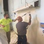 One man spraying paint onto a production facility wall while a second man back rolls the paint that has just been sprayed