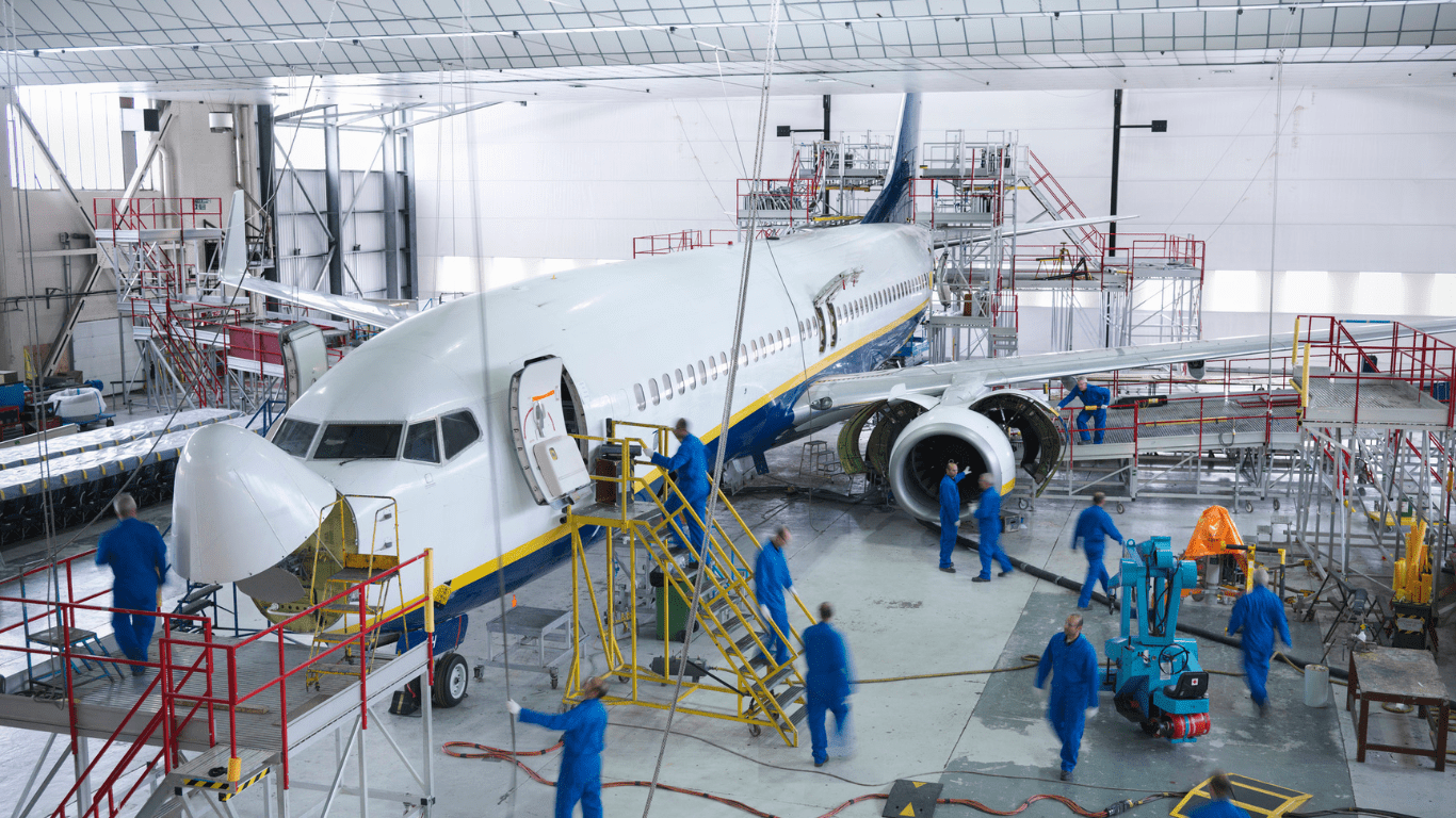The interior of an aircraft assembly facility. A small commercial airplane is worked on by several technicians in blue jumpsuits. Red and yellow metal railings can be seen throughout. There is a large white overhead door in the background.