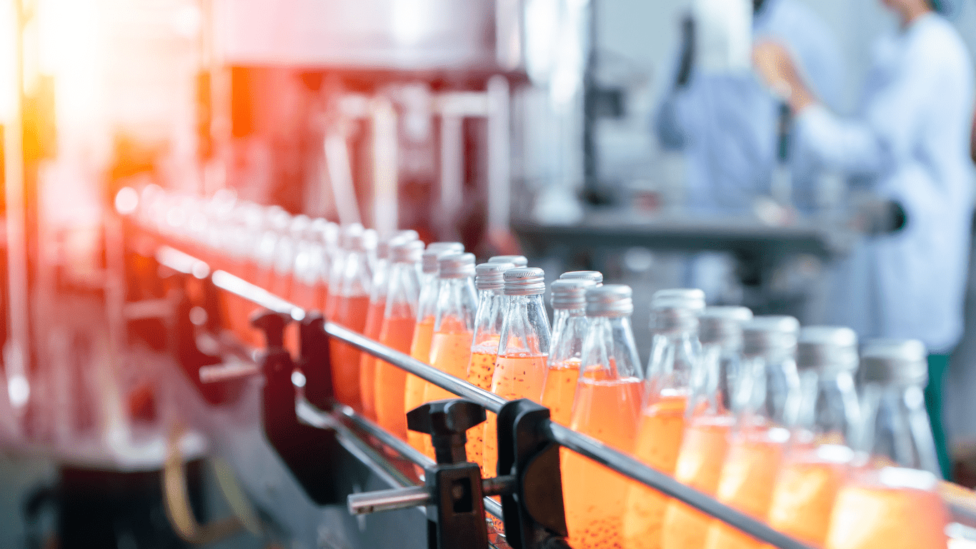 A shot of glass bottles of orange liquid with silver lids traveling down a silver steel conveyor belt. A plant worker can be seen out of focus in the background