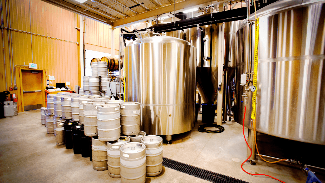 Inside of a commercial brewery with tan metal walls and a tan steel man door to the left. several silver steel tanks can be seen as well as several metal beer kegs stacked 2 high.