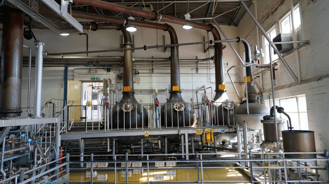 The interior of a commercial alcohol and spirits distillery. 3 large black metal tanks can be seen in the background with pipes connected to them. Everything is surrounded with metal railings. The brick walls are painted white