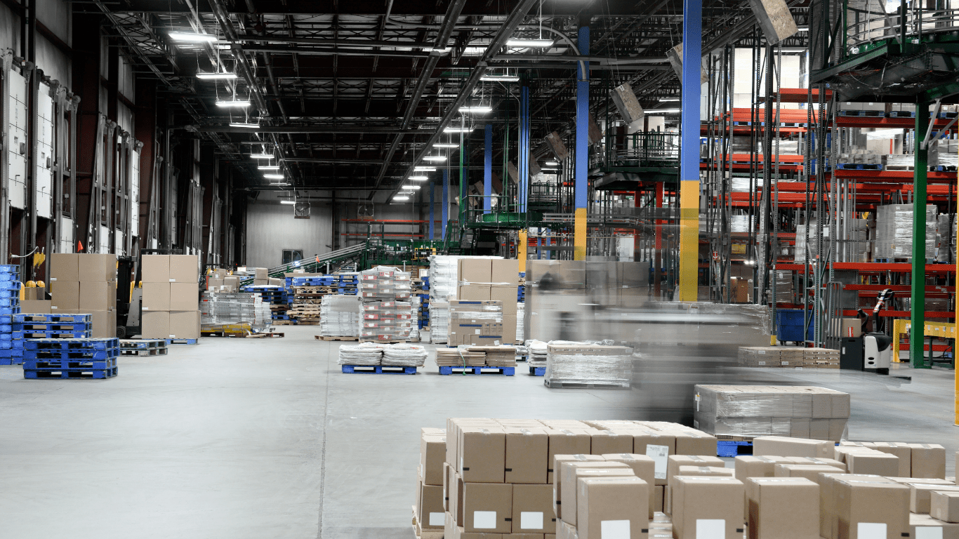 The inside of a commercial distribution center with gray concrete floors, gray metal walls, white overhead doors, and black structural steel and open beam ceilings. Gray and red racking can be seen to the right and several pallets of boxes are scattered throughout
