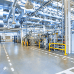 The interior of an industrial manufacturing facility. A forklift lane is marked out with white paint in the middle and walking lanes are indicated with dashed lines on either sides. There are several yellow bollards and railings. All of the visible structural steel is painted white