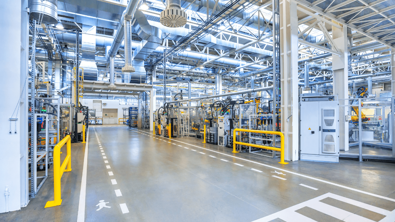 The interior of an industrial manufacturing facility. A forklift lane is marked out with white paint in the middle and walking lanes are indicated with dashed lines on either sides. There are several yellow bollards and railings. All of the visible structural steel is painted white