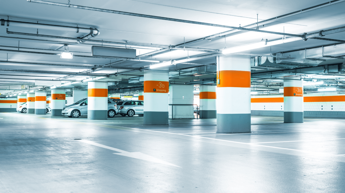The interior of a commercial parking garage with white walls, ceilings, and columns. There are orange stripes throughout. Several silver hatchback vehicles are parked in the background