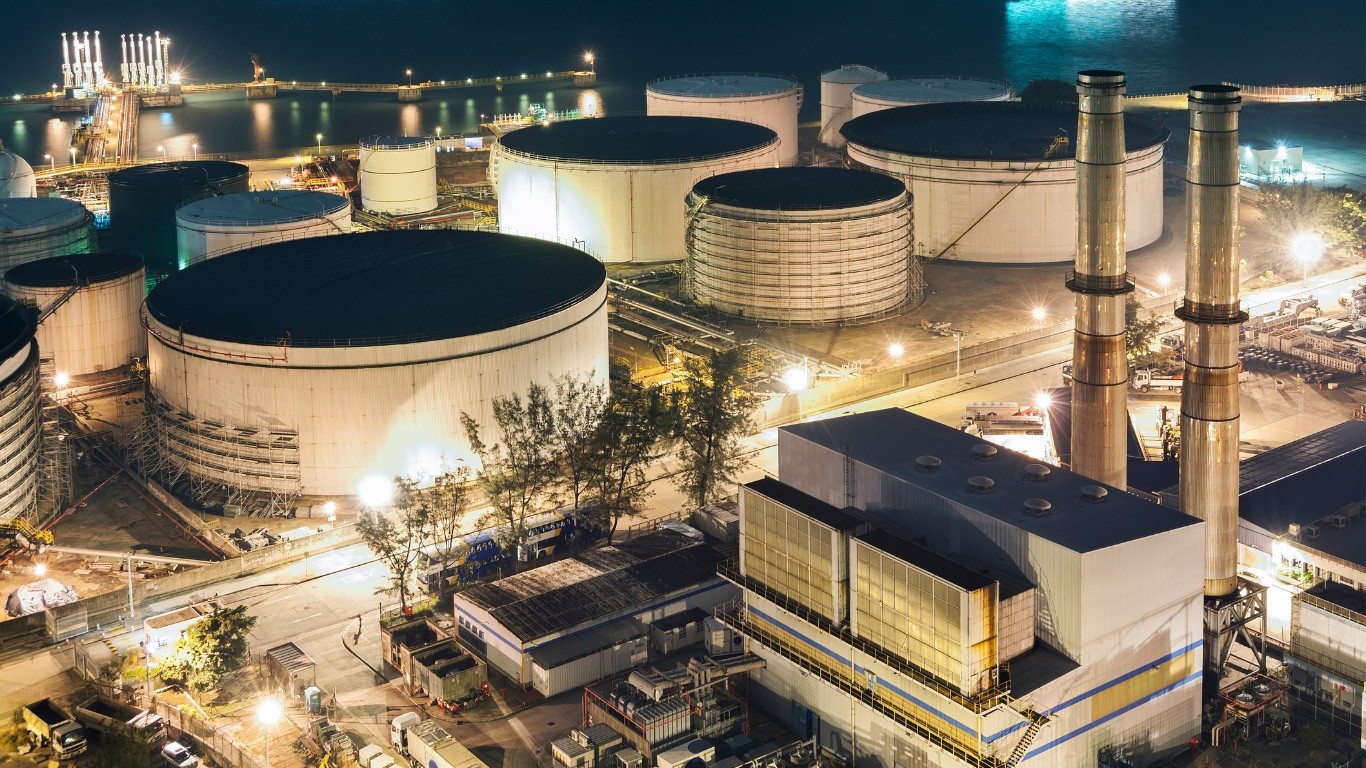 An arial view of a power generating plant. Several cooling towers and smokestacks can be seen. Water is in the background. There is a gray metal building with blue stripes in the foreground. Several vehicles can be seen throughout the complex.