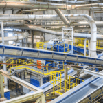 The interior of an industrial processing facility. There are several catwalks with yellow metal railings. There are several chutes and conveyor belts painted blue. There is lots of ductwork in the background