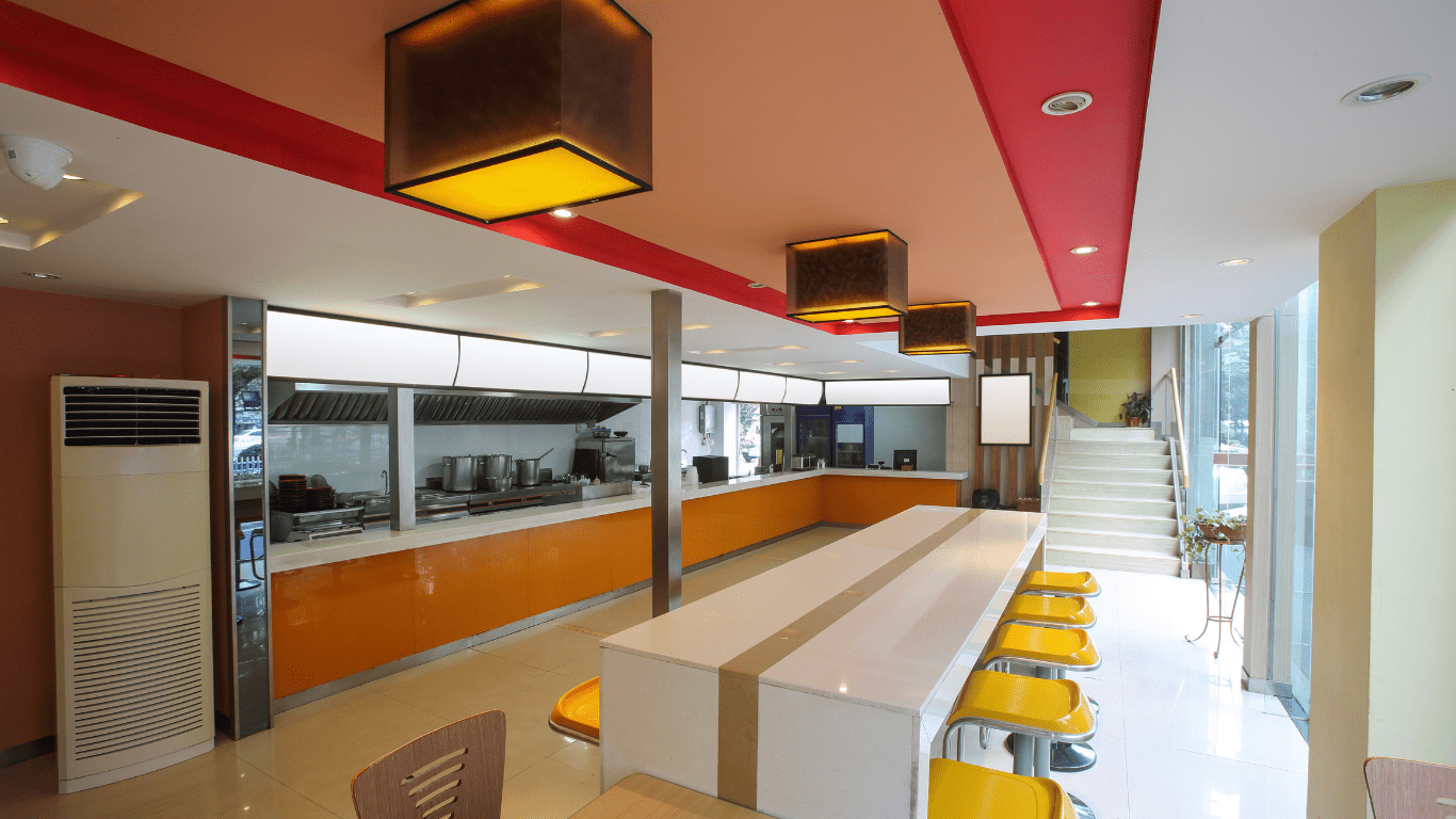 The interior of a fast food quick service restaurant. The walls are painted light green and orange. The ceiling is red and orange and white. There is a long white counter height table with yellow stools on both sides and brown lights on the ceiling.