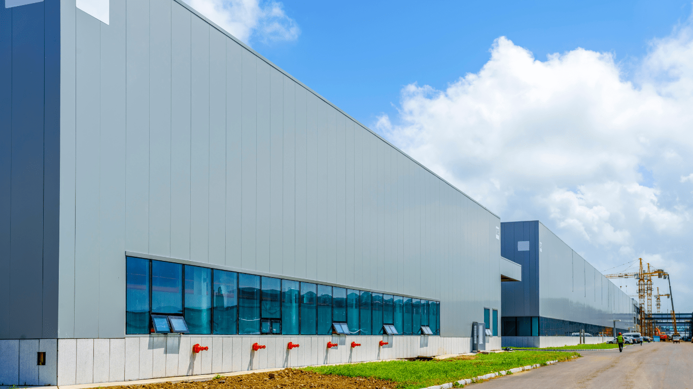 The exterior of a gray metal industrial or commercial building continuing off into the distance where cranes can be seen.