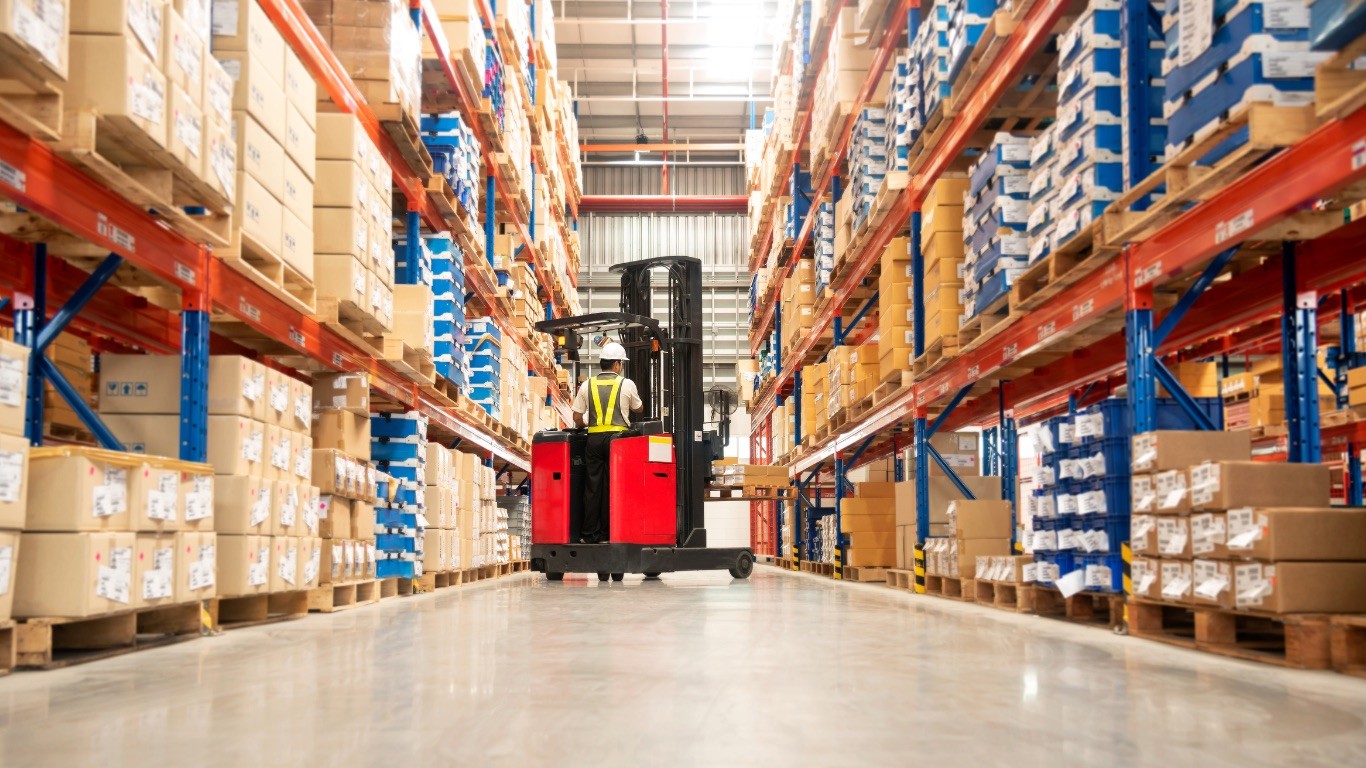 A red and black forklift moves a pallet of boxes down a warehouse aisle with racking painted blue and red stacked high with pallets of boxes