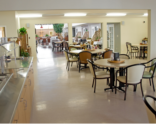 The buffet style cafeteria in a retirement home. Round tables are scattered throughout surrounded by three or four chairs each. There is a stainless steel salad bar to the left. The walls are pale yellow. In the background there are doors that open to n outdoor patio with more tables and chairs.