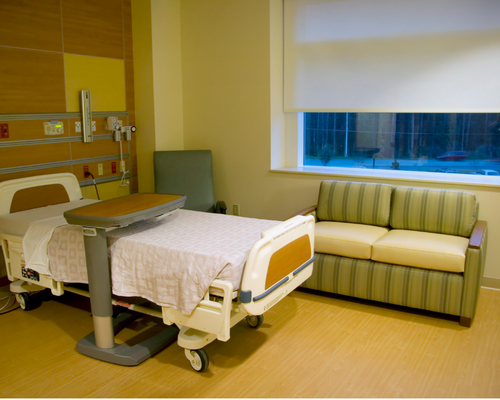 A hospital bed sits in a room in a medical or assisted living facility. There is a gray and brown wood tray table across the middle of the bed, the sheets are white. There is a green striped couch under a partially shaded window against the back wall which is painted yellow. The floor is brown wood