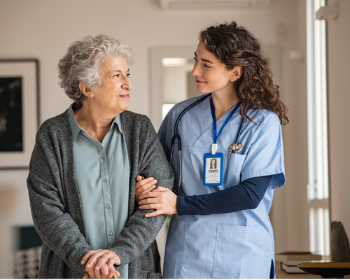 A nurse with brown, curly hair and blue scrubs walks arm-in-arm with a woman with short gray hair and a gray jacket using a cane. They are walking down a cream colored hallway in a freshly painted assisted living facility.