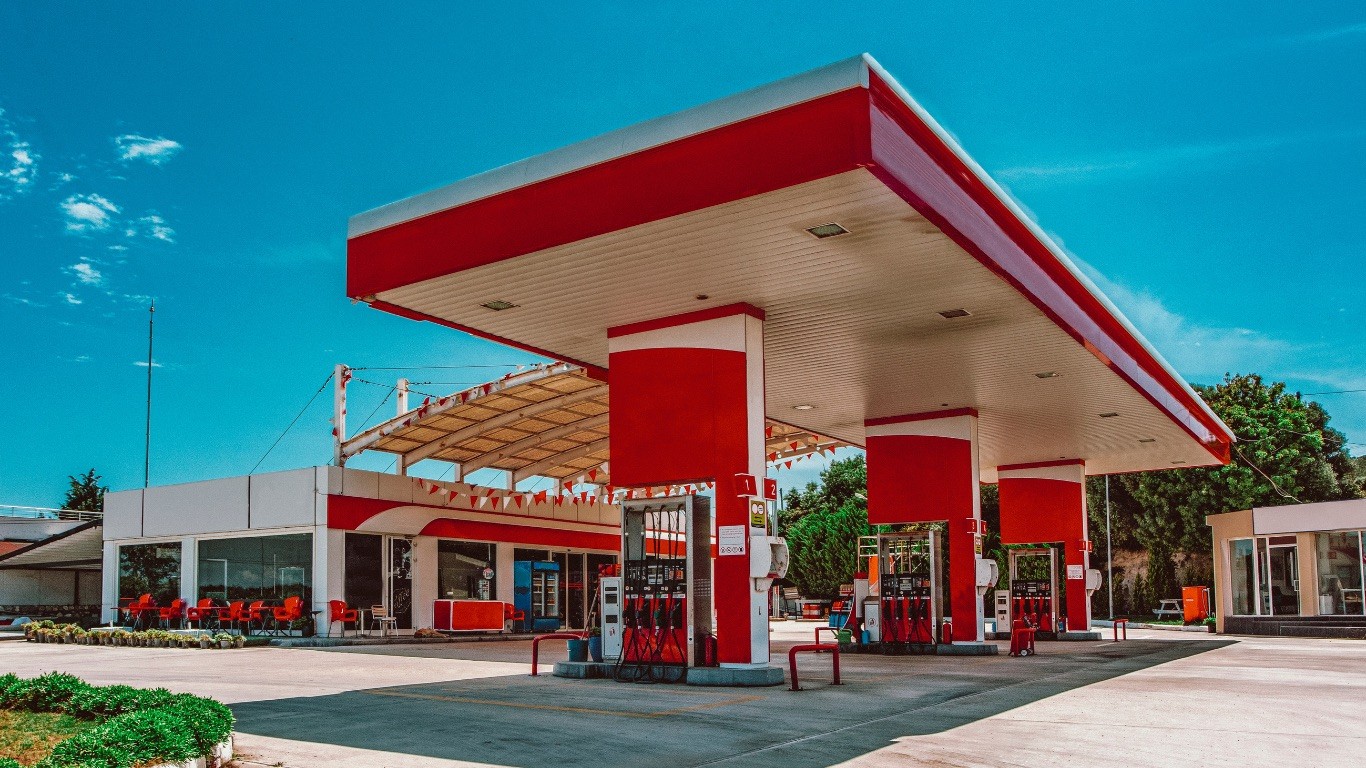 A gas station with 3 pumps and a large canopy painted red and white attached to a white and red convenience store with red chairs outside.
