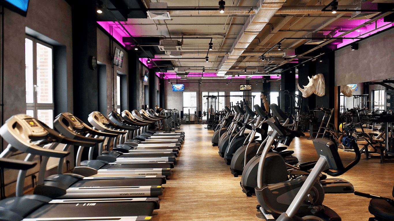 The interior of a commercial gym and fitness center full of treadmills, ellipticals, and stationary bikes. The walls are black and dark gray and the ceiling is off white with visible pipes and duct work. In the background a large white rhino bust is mounted to the wall and someone runs on an elliptical