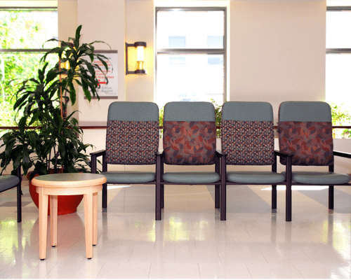 police station painting (3) 4 chairs lined up arm to arm in a beige waiting room with white tile floors. There is a large plant on the left behind a small tan table.