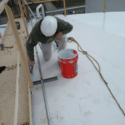 A professional painter in a green shirt, wearing a white hard hat applies white urethane paint to the top of a large white industrial holding tank